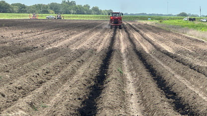 A red tractor drives down long rows of freshly tilled dark brown soil in an open farm field under a clear blue sky.